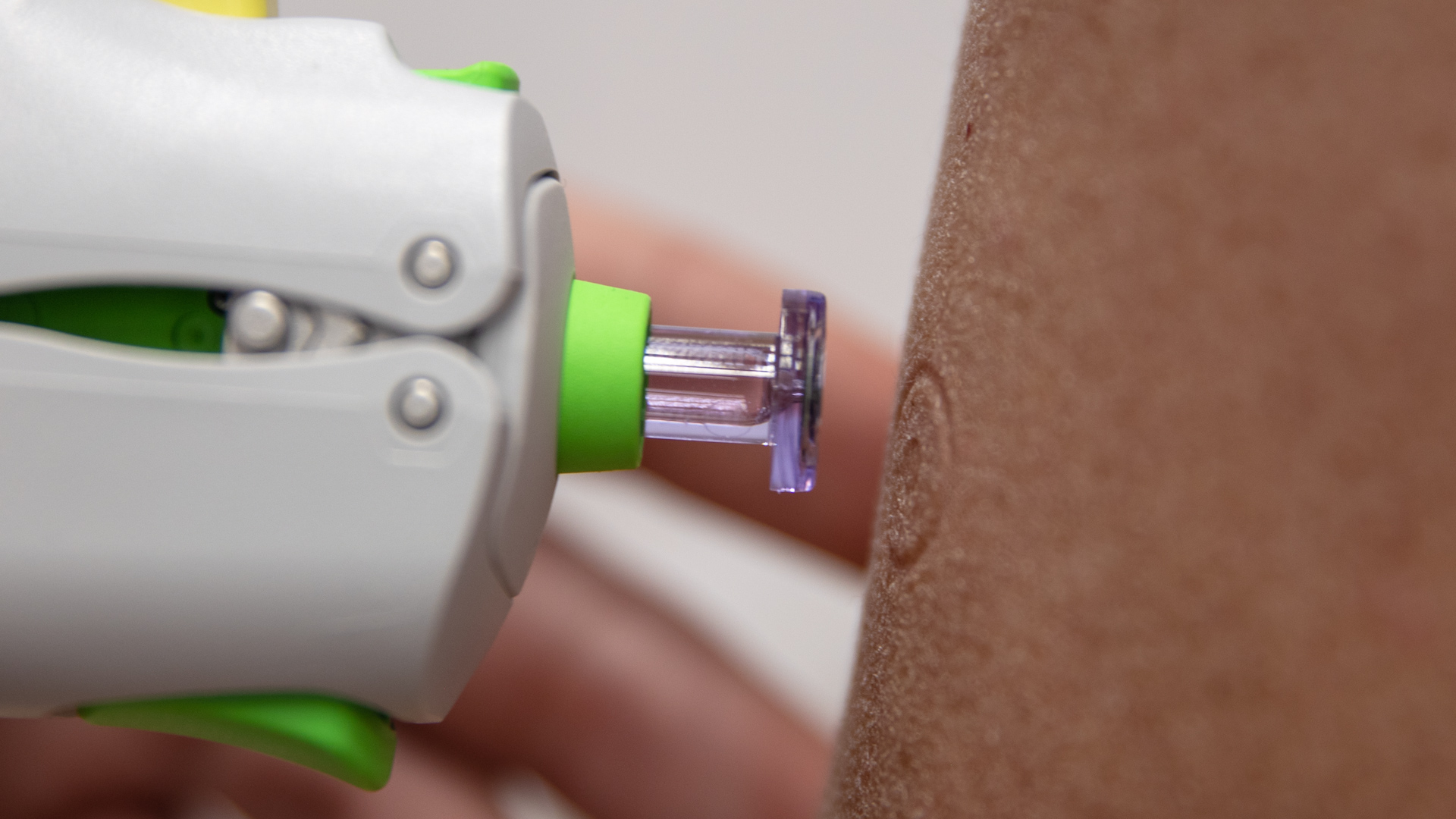 close up of microfluid vaccine delivery to a patient's arm; the device is white with a bright green tip from which protrudes a clear plastic circular end, there's a matching circular mark on the patient's skin